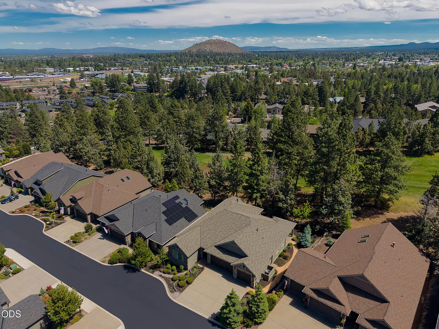 Aerial wide view of northwest Bend with Pilot Butte rising on the horizon, the property's neighborhood held between mature pines in the foreground and the High Desert mountains in the distance