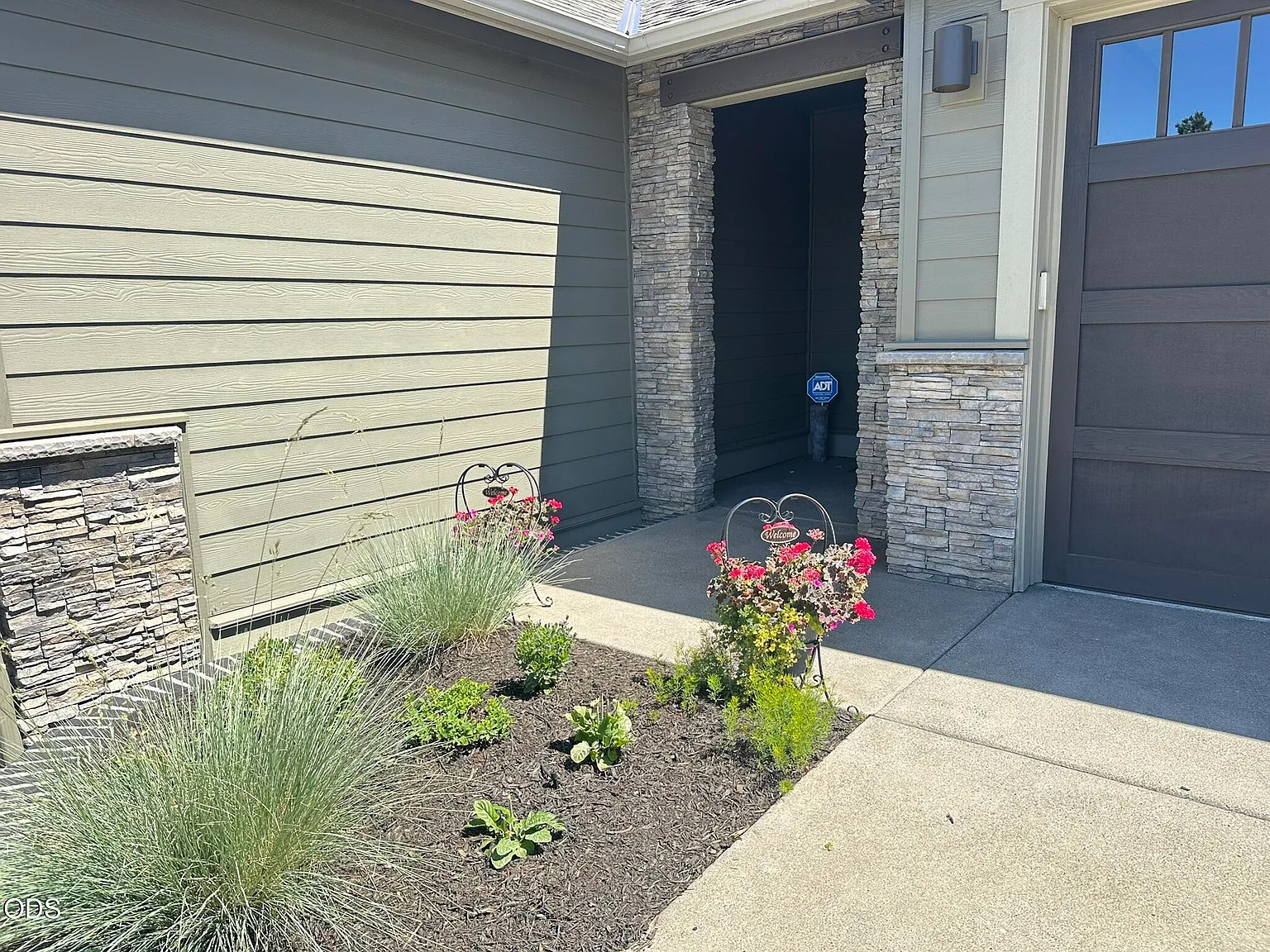 Detail of the covered front entry, showing the stone column at the threshold, dark siding, paneled garage door beside it, and the planted bed of low grasses and flowering perennials in the foreground
