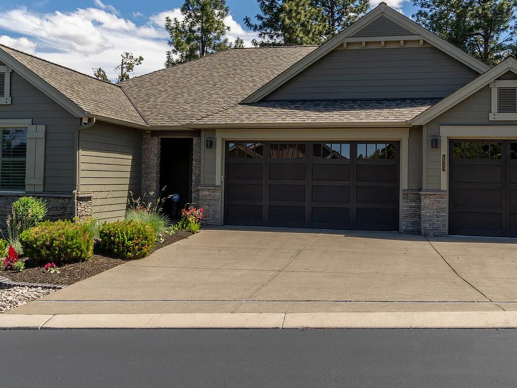 Tight architectural detail of the front facade showing the stacked-stone column base, the dark sage board-and-batten siding, the wood-paneled garage doors, and the front entry framed by low landscaping