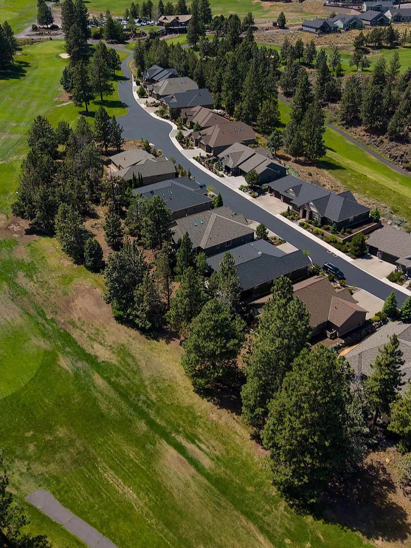 Aerial portrait view of the cul-de-sac, with the row of single-level Craftsman homes lined along the cart path, the fairway grass running parallel, and dense ponderosa pines flanking the green corridor
